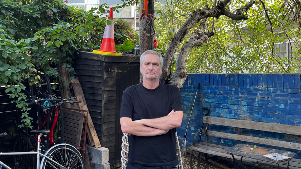 A person wearing a black T-shirt stands with arms crossed in a leafy garden, with bicycles, a wooden bench and a blue brick wall behind.