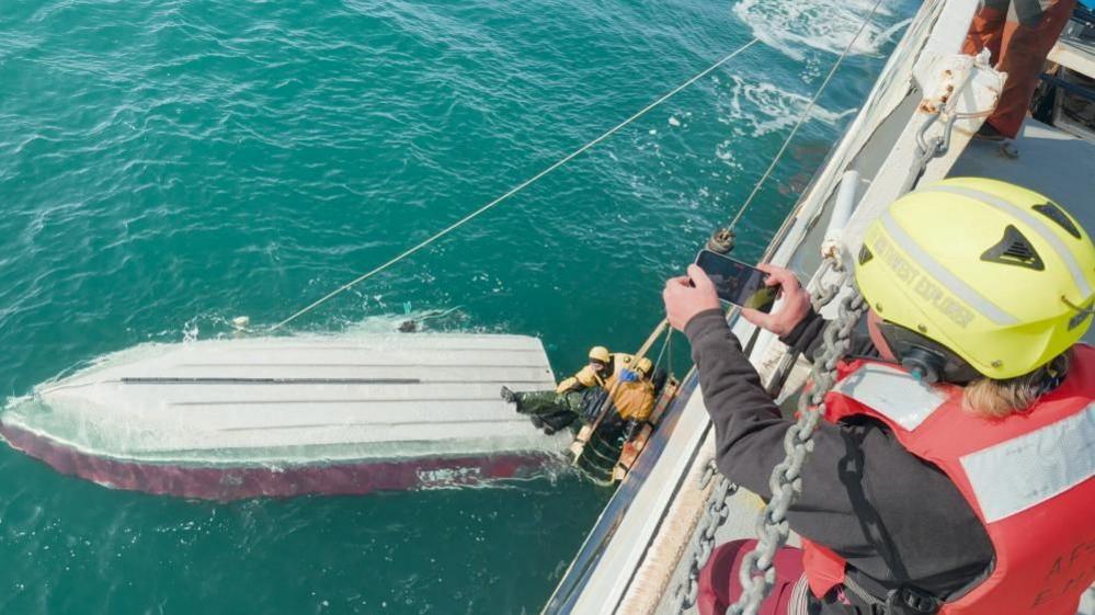 Rescuers aboard a large vessel pulling a teen boy to safety using a pulley system from an overturned boat floating in blue-green waters.