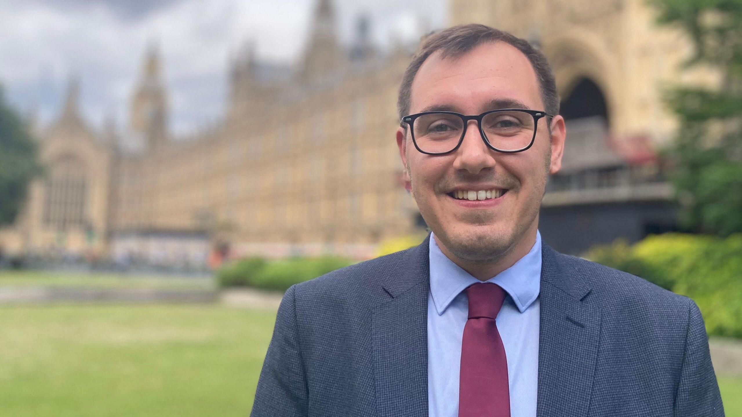 Image shows a man standing in front of the Houses of Parliament in London. He is wearing glasses and has a dark blue suit, with a light blue shirt and red tie.