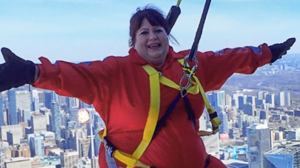 Image shows Julie Fadden abseiling down the side of a skyscraper in Seattle. She is wearing a red jumpsuit and the Seattle skyline can be seen behind her.