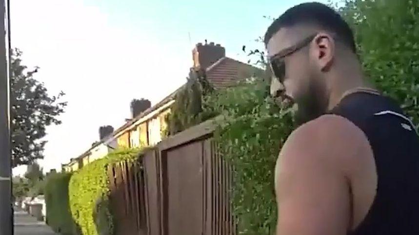 Screengrab of video footage showing Dogan's side. He is wearing a black tank top, short blak hair, a beard and sunglasses. He is positioned to the right of the image with the rest showing a pavement and the side of about five houses.