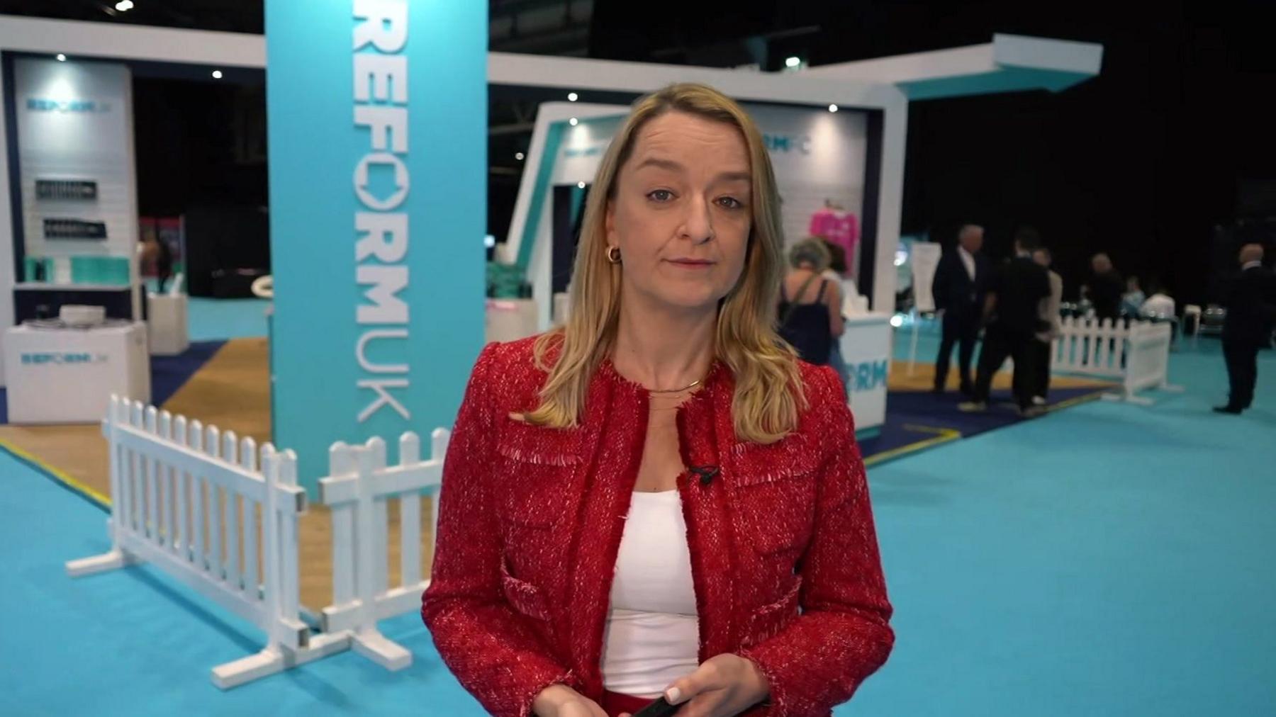 Laura Kuenssberg looks at the camera in a red jacket, in front of light blue Reform banners and a light blue carpet.