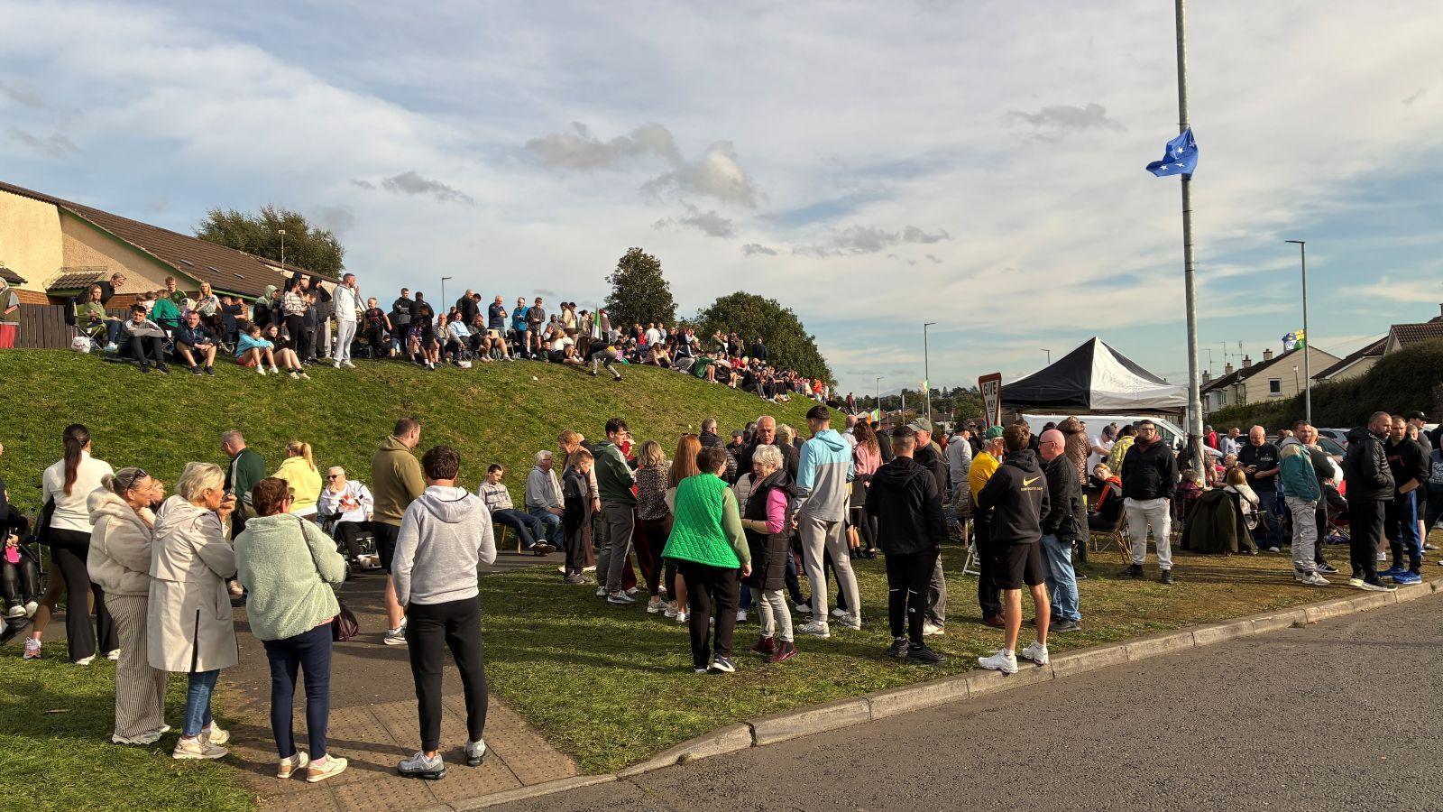 A group of protesters standing on the grassy bank at the side of the road. There are about 100 people seen in the picture wearing a variation of clothing.