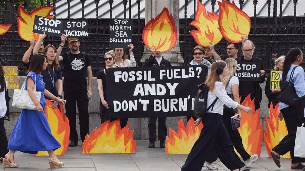 Climate activists stand outside a wrought-iron fence holding flame-shaped placards and signs opposing North Sea oil and gas projects, including Rosebank and Jackdaw, while pedestrians walk past in the foreground.