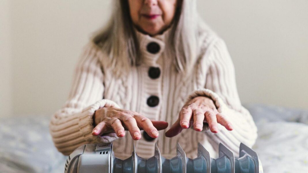 What is the triple lock and why are people talking about it? An older woman wearing a thick cream cardigan sits on a bed holding her hands over an electric radiator.