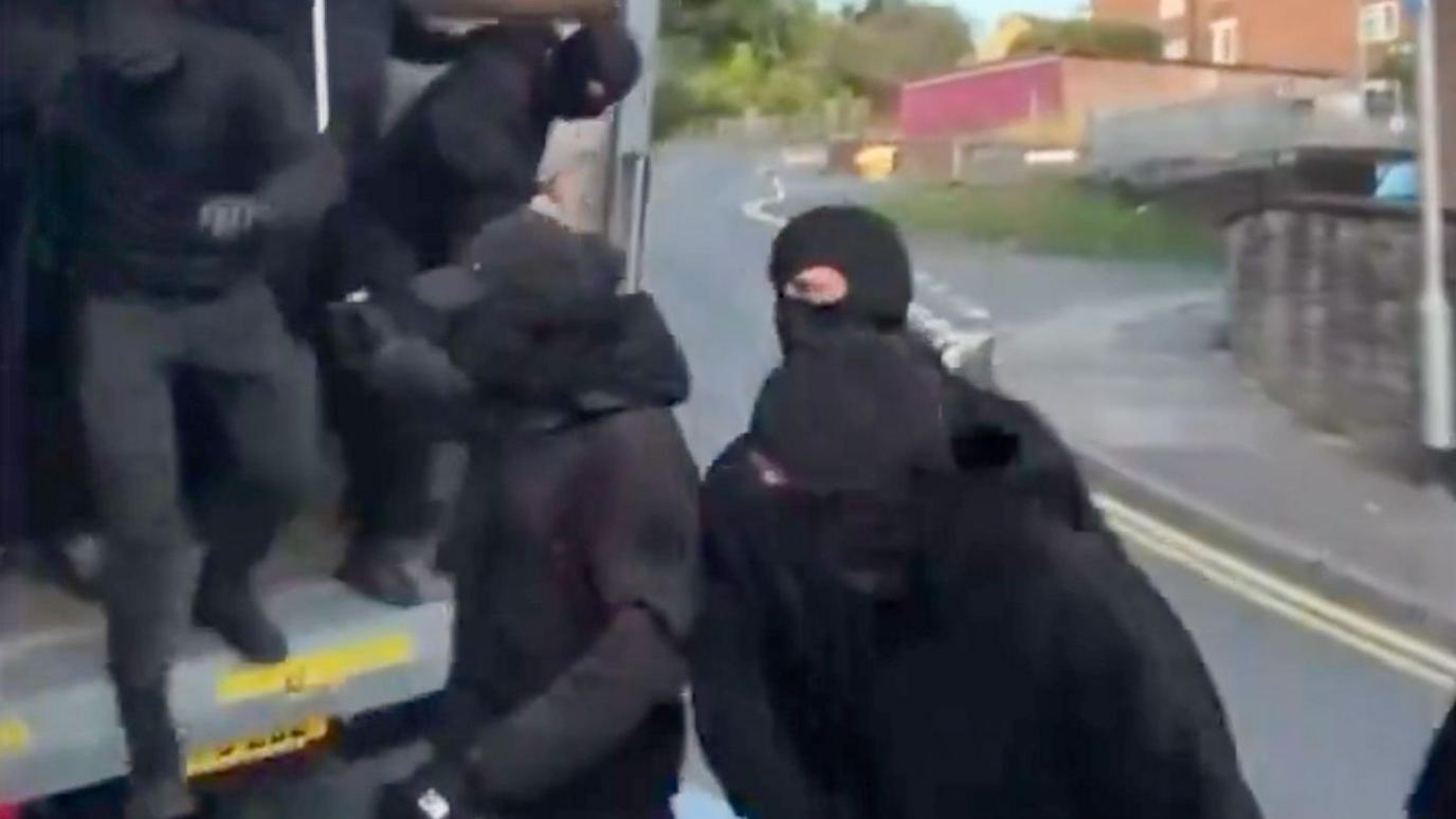 Men in black and balaclavas getting out of the back of a lorry.