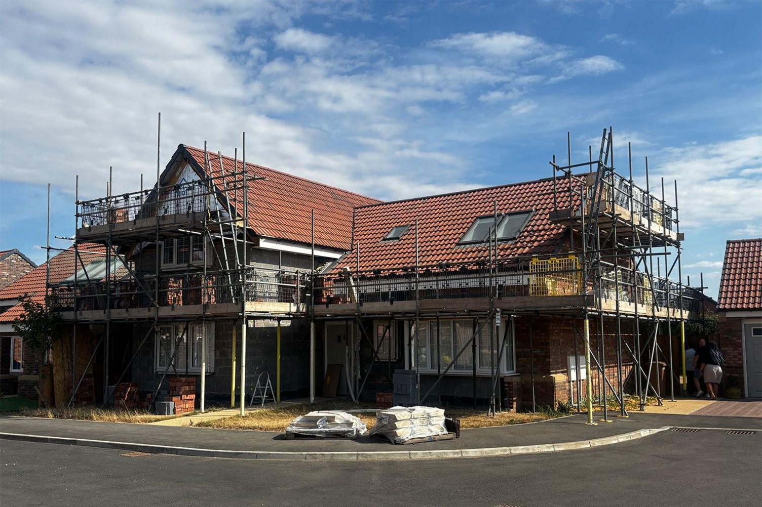 A two‑storey house stands surrounded by scaffolding, with red roof tiles and skylights visible. Construction materials, including wrapped supplies and stacks of bricks, sit in front of the partially finished exterior under a cloudy daytime sky.