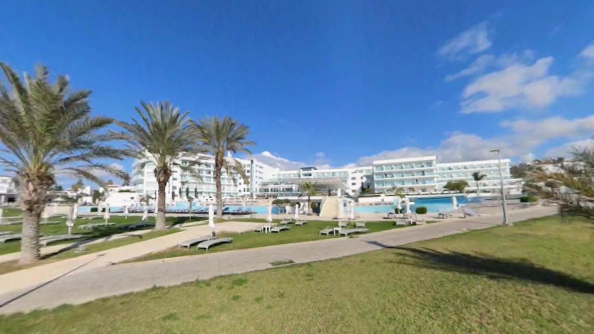 A white hotel complex, with palm trees in the near left distance and a public path - there is a swimming pool just in view in the foreground