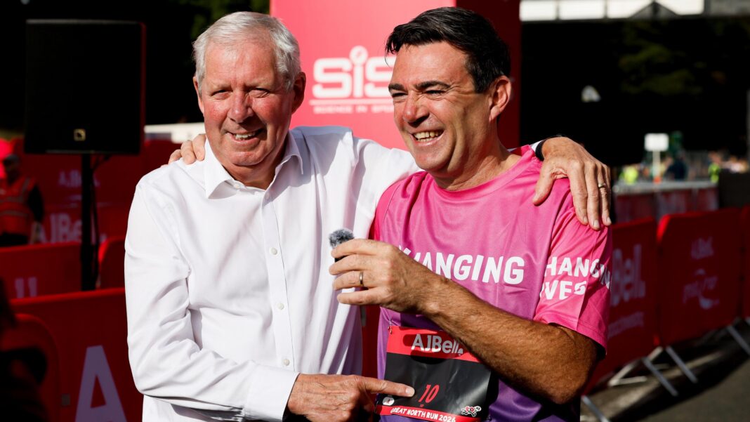 Brendan Foster, who has short white hair and is wearing a white shirt, has his arm slung over Andy Burnham who is wearing a pink running shirt with the number 10 pinned to it. They are both smiling brightly. Burnham, who has short black hair, is also holding a small clip on microphone.