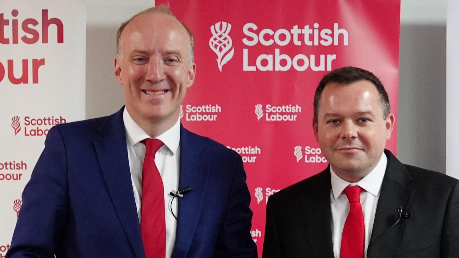 Two men in suits with matching red ties pose in front of a red-and-white backdrop displaying the Scottish Labour logo and name.