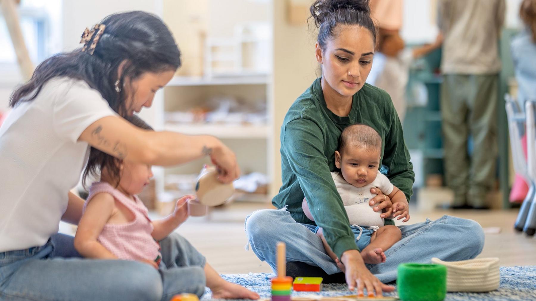 Two women and two young children sitting on the floor in a playroom. One woman holds a baby while reaching towards a set of colourful toys on the rug. The other woman sits beside a toddler who is playing with wooden toys. Shelving with baskets and play materials is visible in the background.