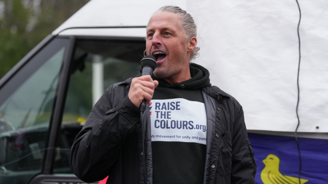 Image shows a man with tied back blonde/grey hair holding a black microphone. He is talking and smiling, wearing a black jacket and a black hoodie with a white logo that says in blue 'Raise The Colours'