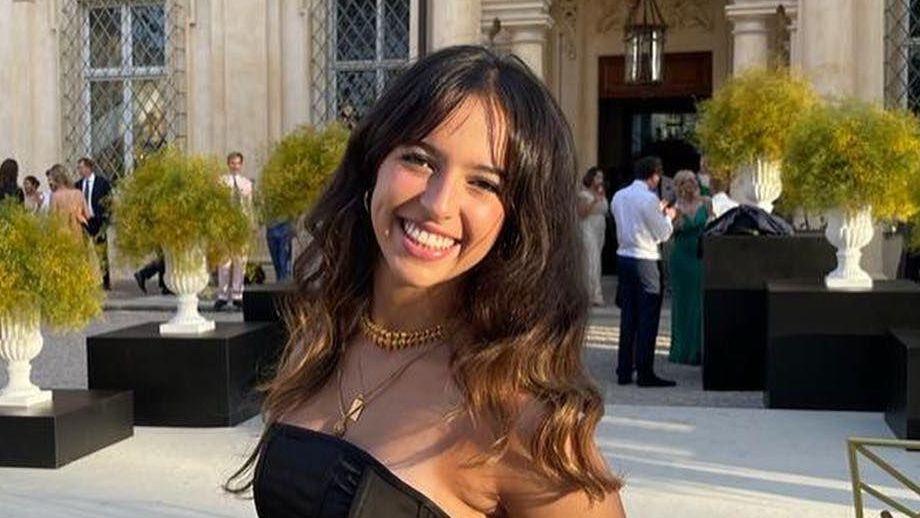 A photo of Grace O'Malley-Kumar, a young mixed-race woman with long black hair. She is wearing gold necklaces and a black strapless dress. She appears to at an event and is standing in front of a large venue, smiling. A number of other guests can be seen in the background.