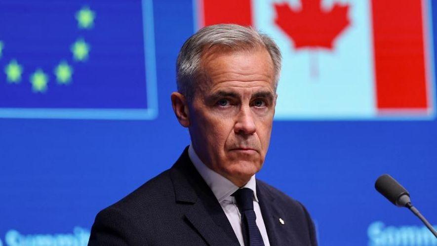 A file photo of Mark Carney, who has grey hair and wears a suit, in front of a screen that shows a digital version of the Canadian and EU flags