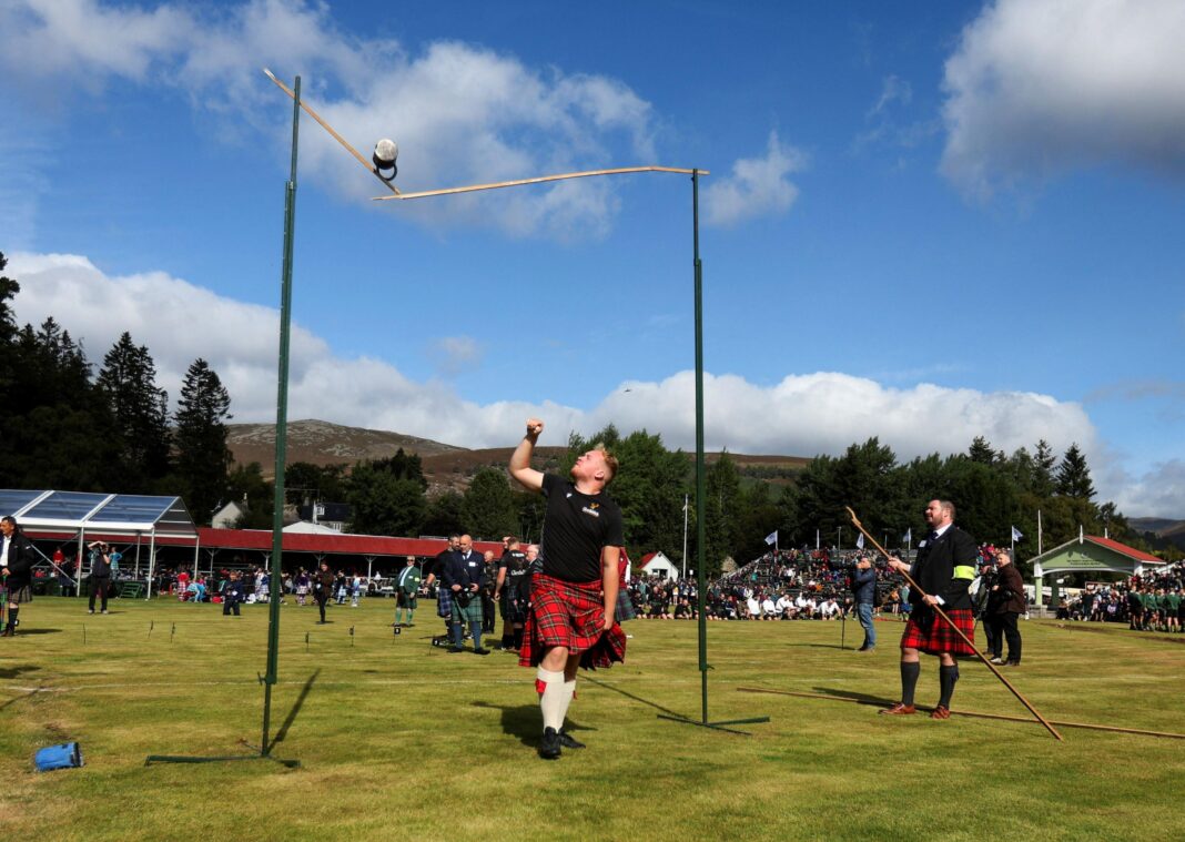 King and Queen join Highland games fans at Braemar Gathering a kilted man looks up as a heavy weight he has thrown lands on and brakes a high bar