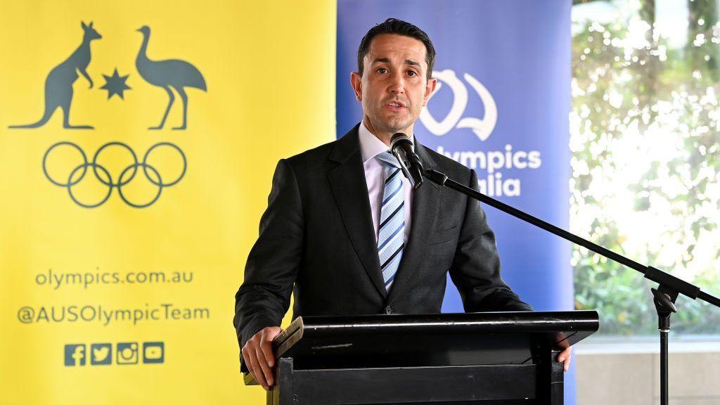 A man in a suit stands at a podium in front of a yellow Olympics banner with a kangaroo and Olympics rings