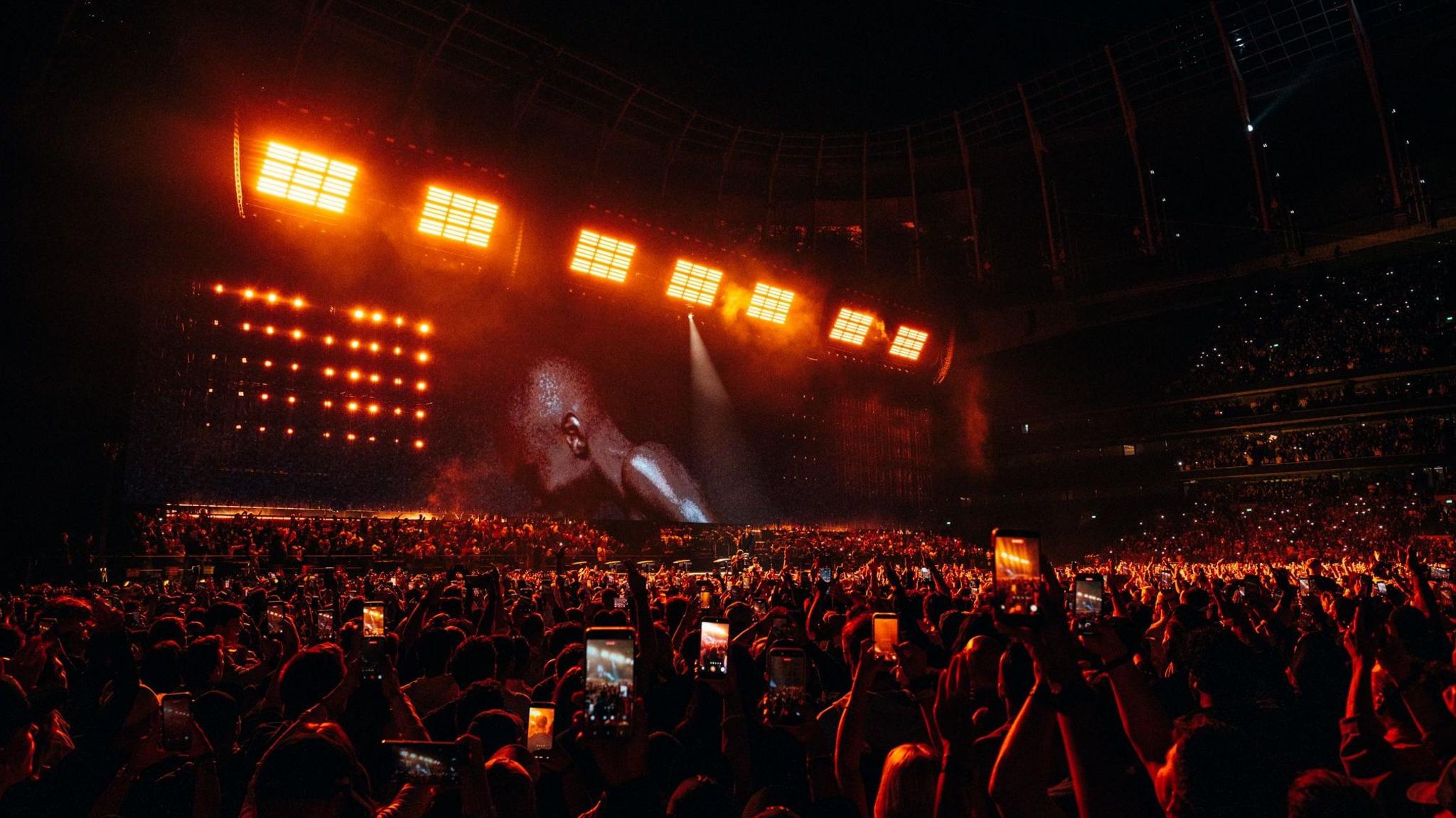Jay-Z performs on stage as a sea of fans watch along. There are phones raised in the air and there is a red hue cast over the crowd.