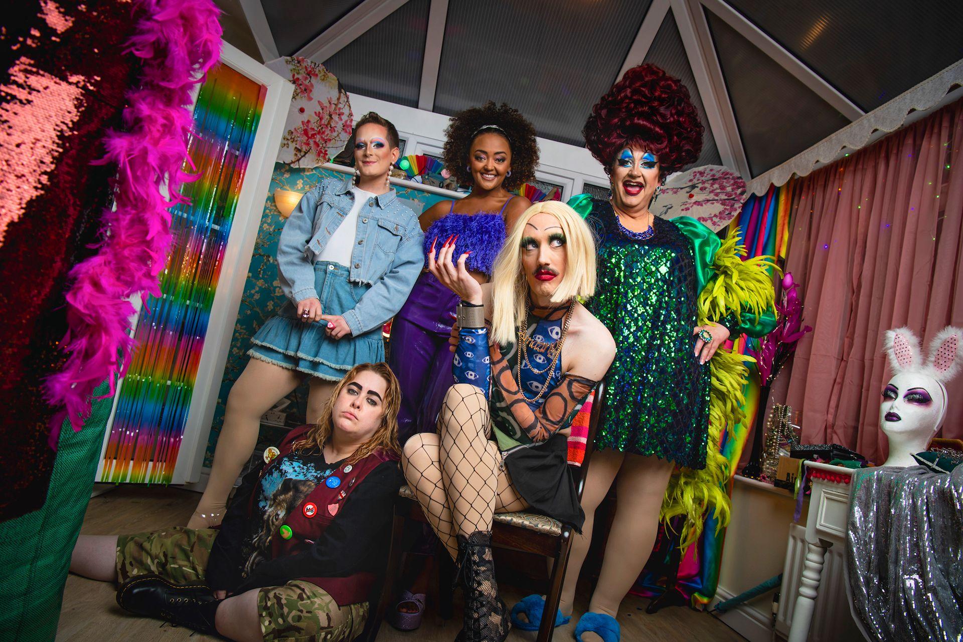 A promotional shot for Smoggie Queens which shows three men in drag posing in a conservatory alongside two women.  In the foreground, one person sits on a chair wearing a blonde wig, fishnet tights, a patterned blue top and black lace-up boots, while another sits on the floor in camouflage shorts, black boots and a burgundy vest with badges. Behind them, three people stand wearing a denim outfit, a purple dress, and a green sequined dress with bright feathered sleeves. The room contains rainbow curtains, pink feather boas, costume accessories, and a white mannequin head wearing bunny ears on a small table to the right.