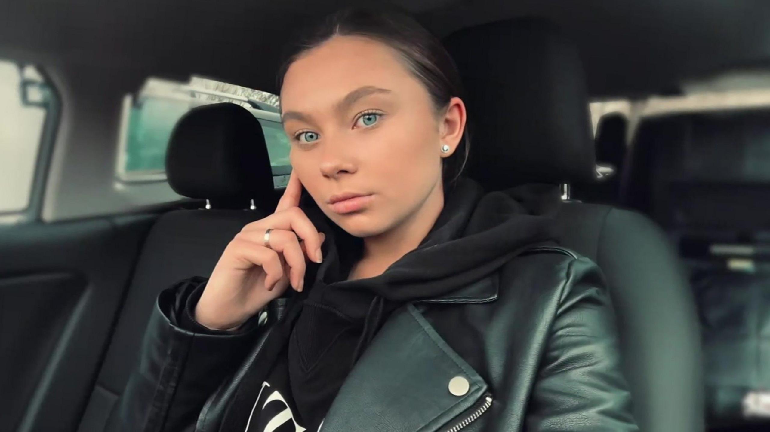 A selfie of Jaysley Beck sitting in a car with a black interior. She is wearing a black hoodie underneath a black leather jacket. She has bright blue eyes and straight brown hair which is pulled back into a ponytail.