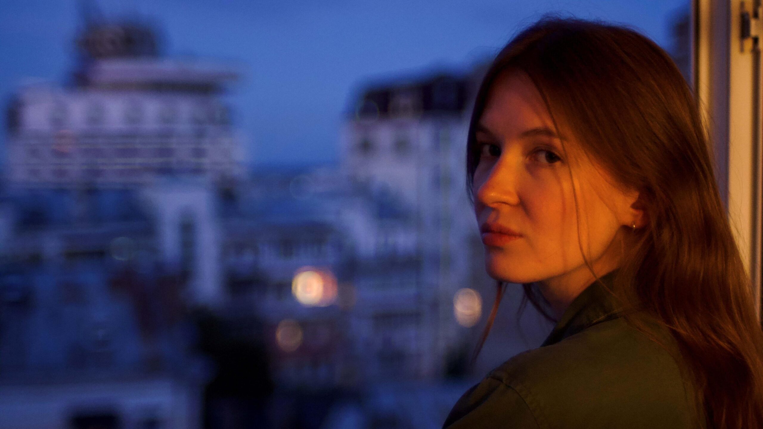 Daria is pictured by the window of an apartment building, with blurred views of the city behind her