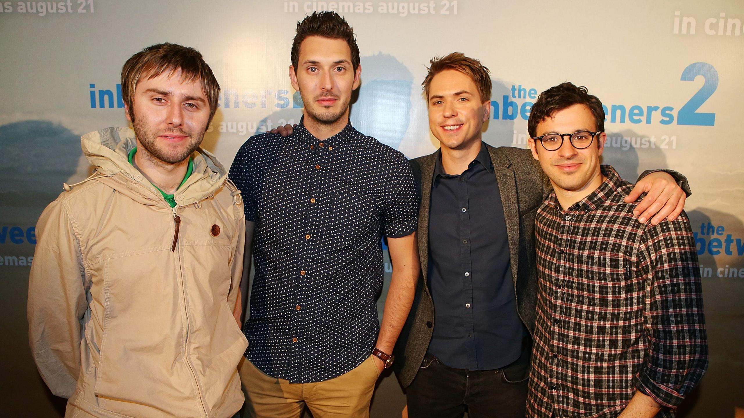 James Buckley, Blake Harrison, Joe Thomas and Simon Bird pose at a premier of The Inbetweeners 2 in front of a billboard of the film. James wears a grey waterproof, Blake blue and white patterned top, Joe wears a grey blazer over a navy shirt and Simon wears checked patterned shirt.