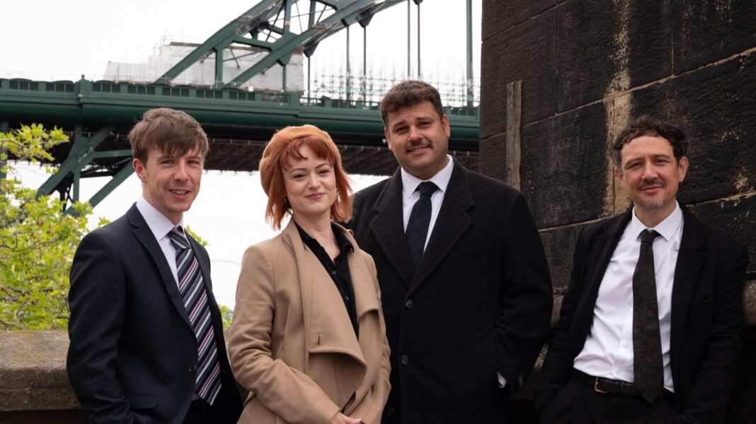 Our Friends in the North still relevant – writer Ryan Nolan, Sam Neale, Jack Robertson and Scott Turnbull stand and smile in front of the Tyne Bridge in Newcastle in costume for a publicity shot for the play. Ryan is wearing a dark suit and multi-coloured tie, Sam a beige coat and beret and black blouse, Jack a dark jacket, white shirt and tie, and Scott a dark jacket white shirt and dark tie.