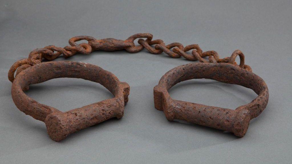 Two iron shackles consisting of iron loops joined together by a length of iron chain. The iron is heavily rusted. These would have been used to restrain enslaved African people forcibly transported to North America and the Caribbean.
