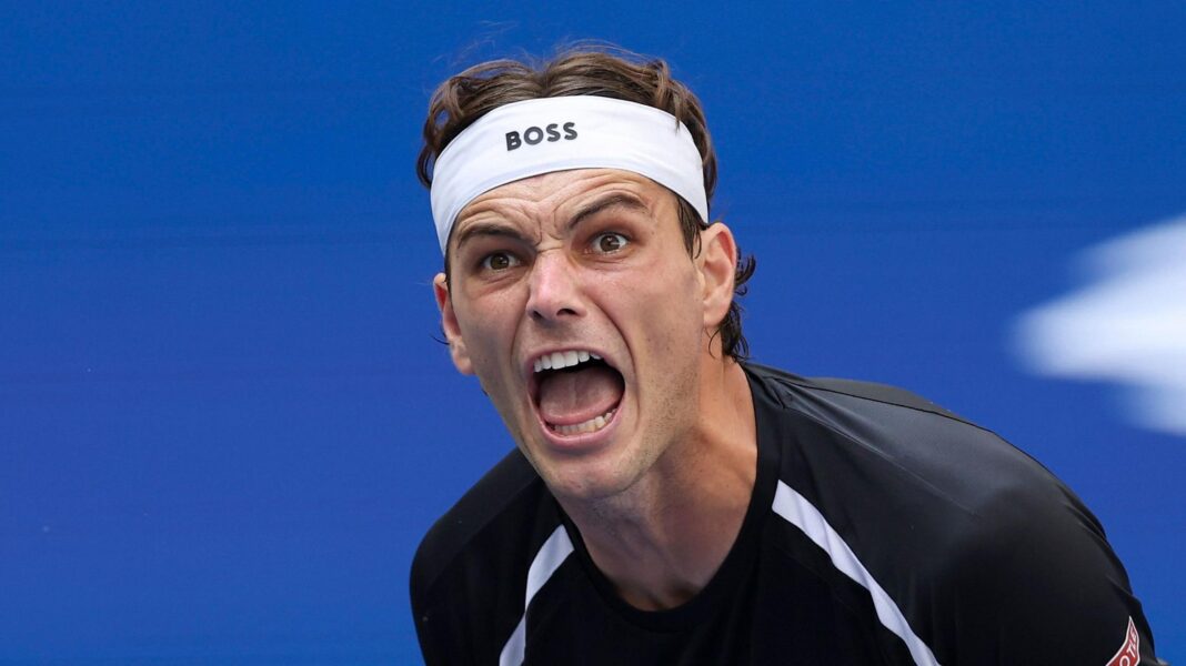 Taylor Fritz, wearing a black-and-white T-shirt and white headband, screams during a US Open match
