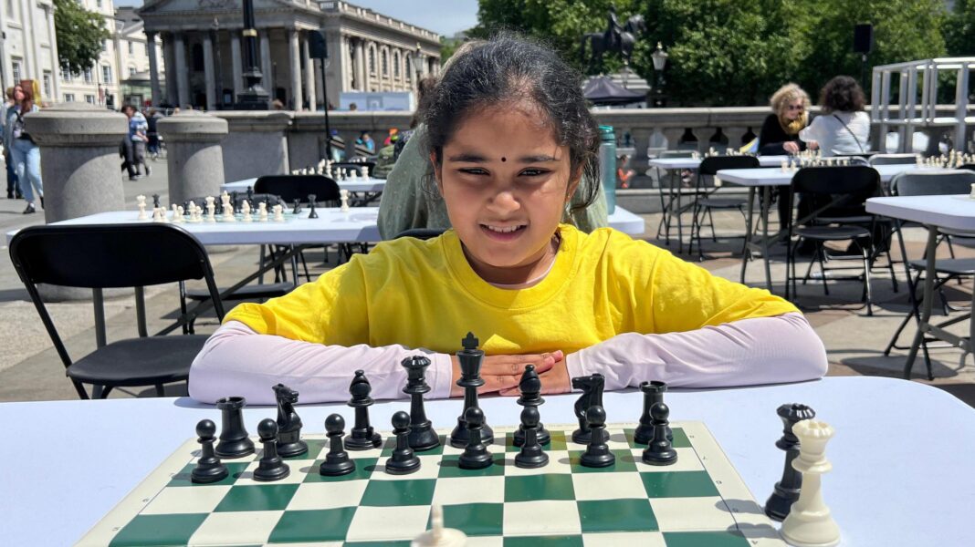 A young girl in a yellow t-shirt sits at a chess board set up on a table in Trafalgar Square in central London. She is facing the camera and is in the middle of a game. There are other tables set up for a chess game in the background.