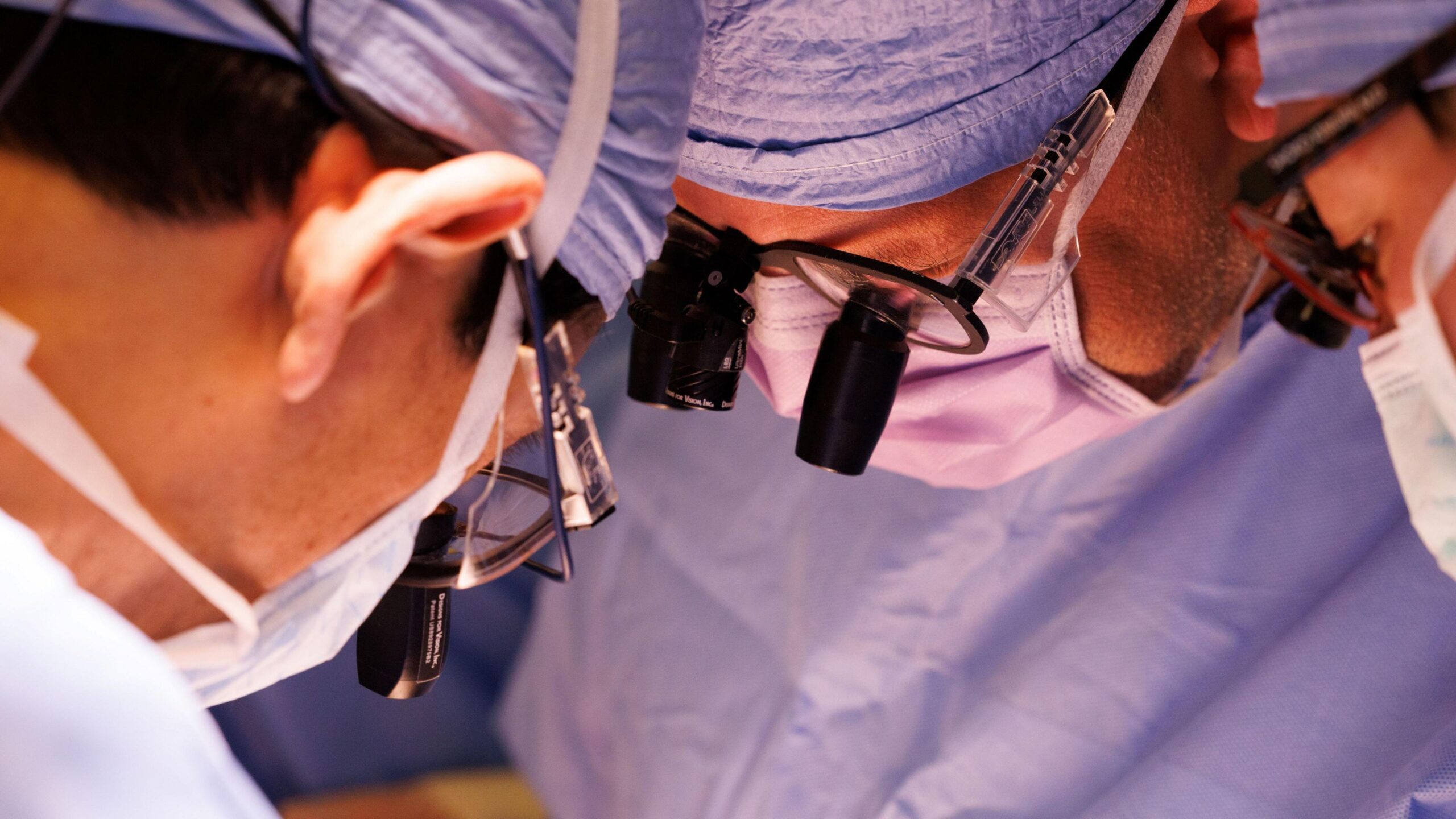 A tightly framed photograph shows several members of a surgical team gathered closely during a procedure. Each person is wearing blue surgical clothing, face masks, and magnifying lenses mounted on protective eyewear. The image focuses on their faces and equipment as they lean inward and look down toward an area outside the frame. Bright operating-room lighting illuminates the scene, while the background remains softly blurred in shades of blue. The composition emphasises concentration, precision, and the collaborative nature of a medical procedure.