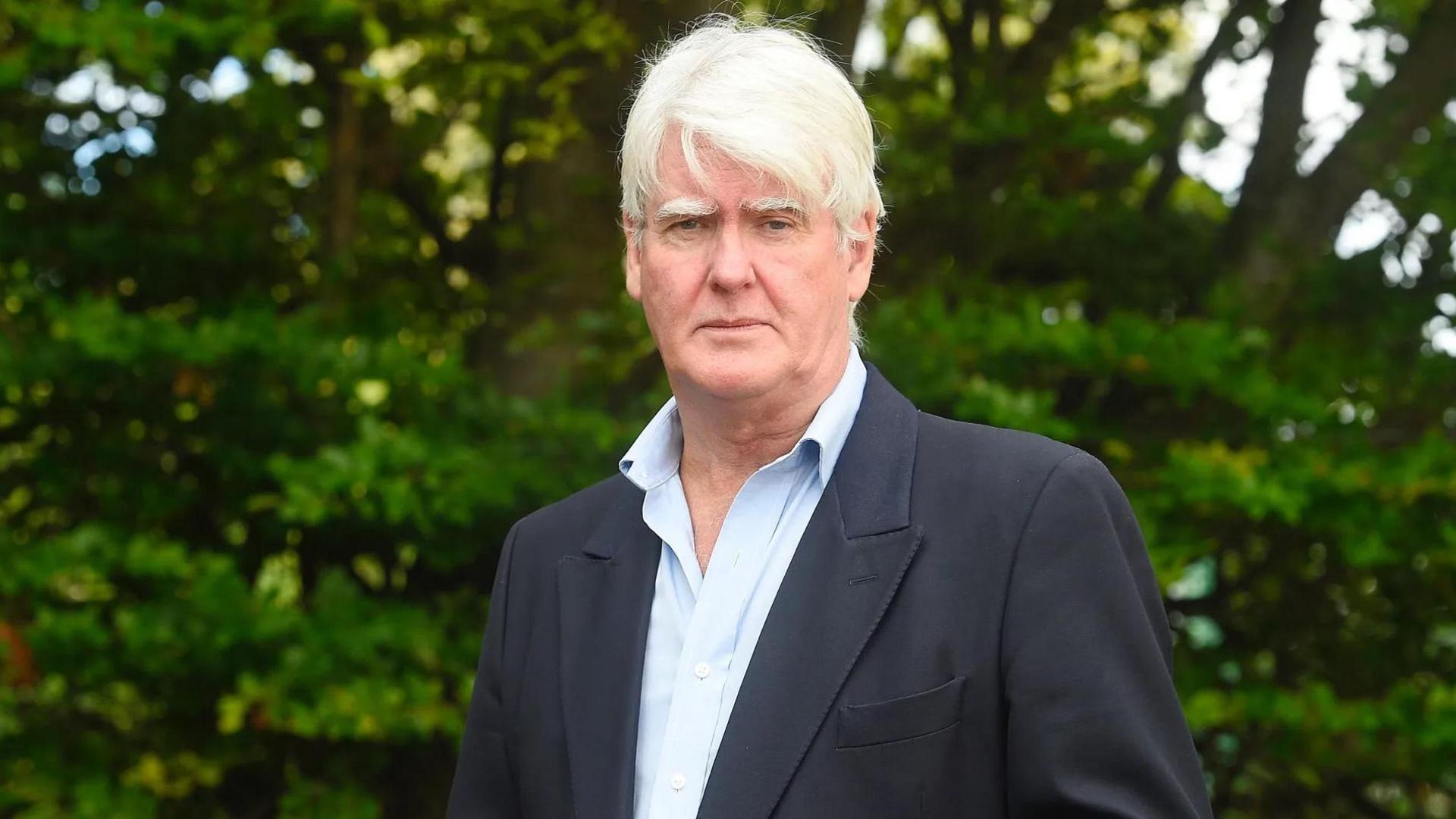Former British Army lieutenant colonel Stuart Crawford wearing a light blue shirt and a blazer. He is surrounded by trees and is looking directly into the camera. His expression is serious.