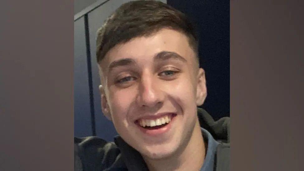 Jay Slater, a young man with brown hair and stubble has a bright smile
