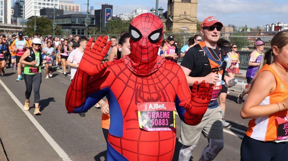 A runner in a spiderman costume with arms raised crossing the Tyne Bridge. To either side and behind are fellow runners.
