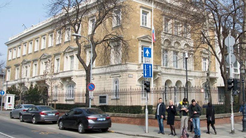 A building in the centre of Budapest surrounded with a metal fence. Five people stand on the pavement at the corner of a road where three cars are seen parked.