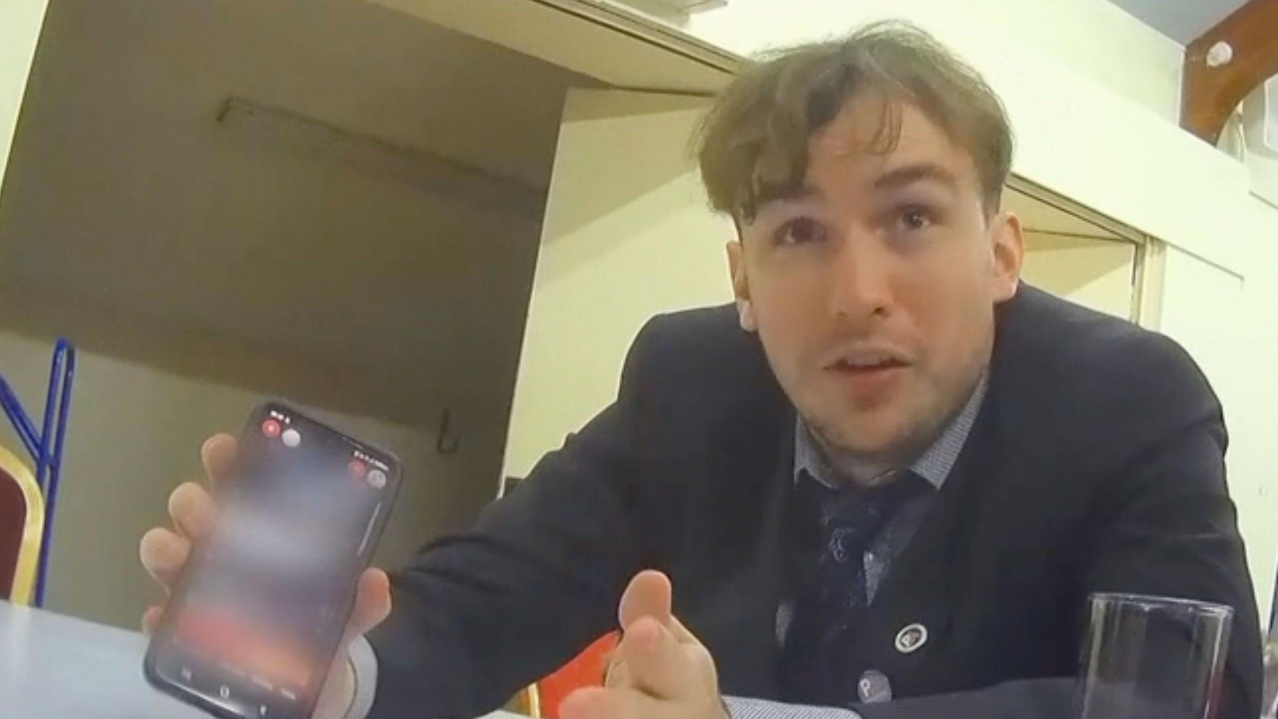 Webster, who is wearing a suit, showing an undercover reporter something on his phone; the screen has been blurred