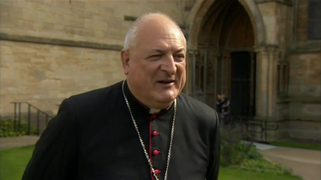 A man looking to the right-hand side of the camera. He is wearing a black cassock and a silver chain around his neck. He's standing in front of a church.
