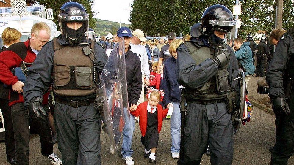 Police officers in riot gear escorting a young child. There is a crowd of people around them.