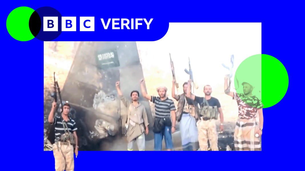 Houthi fighters raise weapons as they stand in front of the tail fin of a crashed Saudi F-15 jet