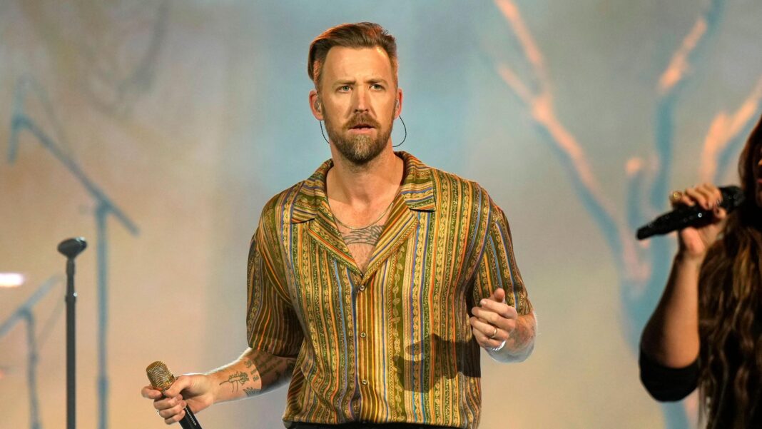 Charles Kelley of Lady A performing onstage during the 2024 People's Choice Country Awards at Grand Ole Opry House in Nashville, Tennessee. He is wearing a gold stripey shirt and holding a microphone.
