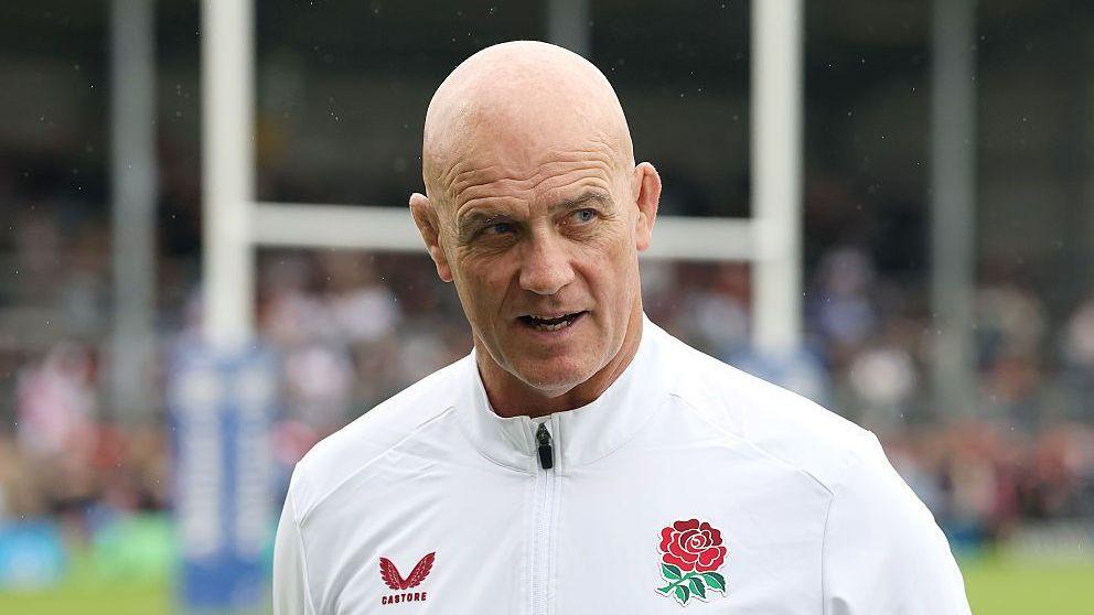 Head and shoulders of John Mitchell, wearing a white England tracksuit top with a red rose on the left breast
