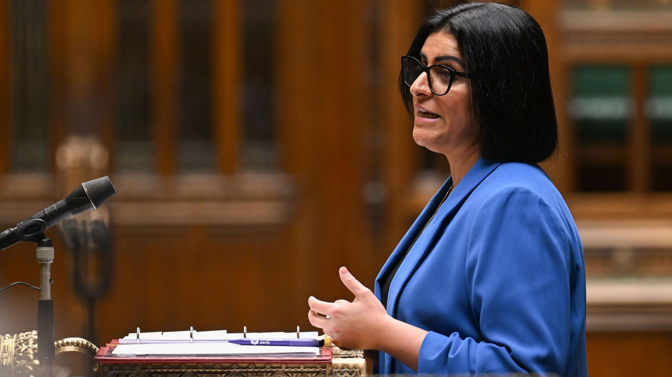 Shabana Mahmood is wearing a bright blue blazer and standing side-on at a microphone beside an lectern with an open ring binder and pen. She has dark hair and large dark-framed glasses. One hand is raised in a speaking gesture. Warm-toned wooden panelling and shelving are blurred in the background.