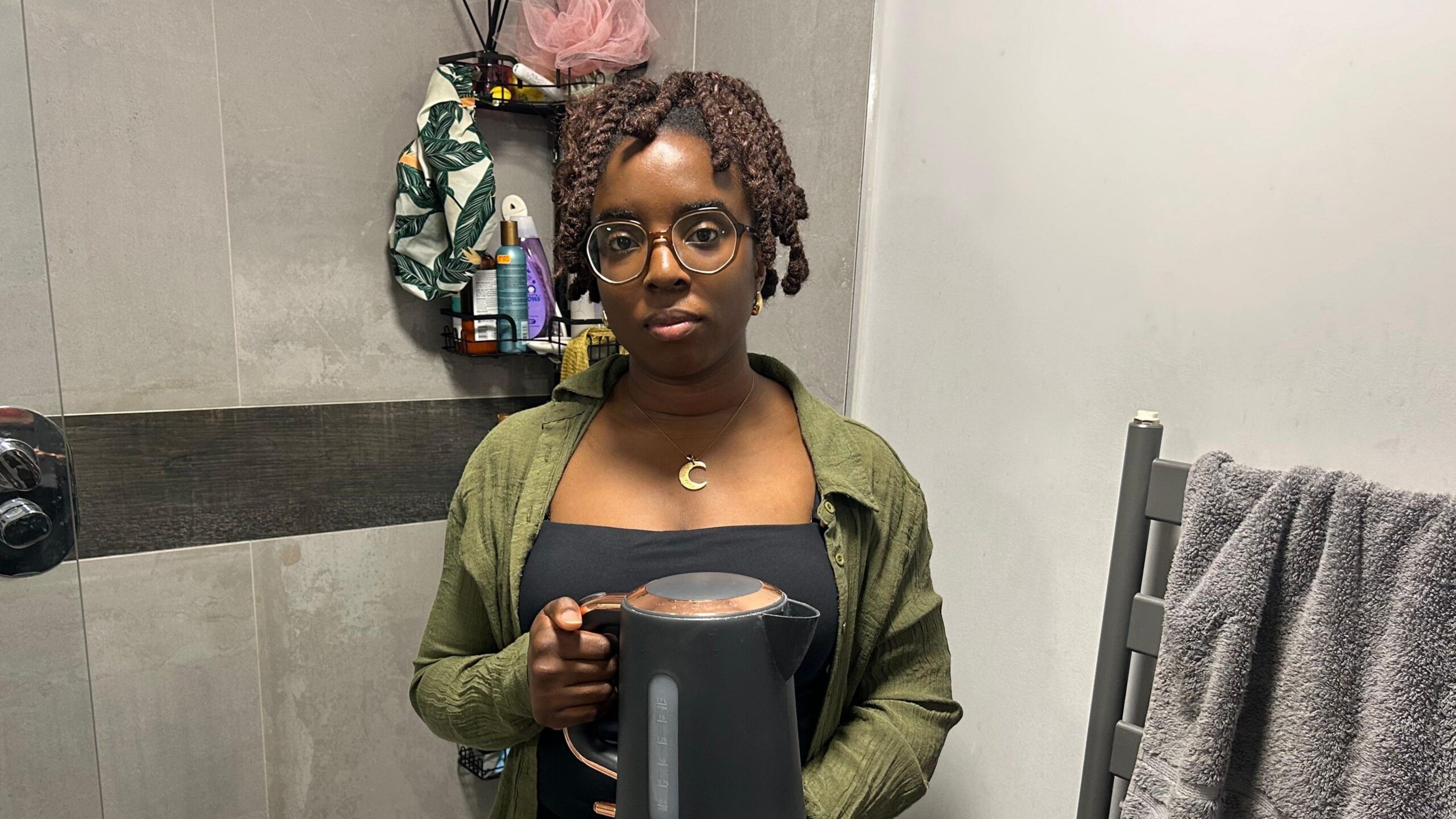 Zainab Ottun holds an electric kettle and is standing in a grey bathroom. She has glasses on and is wearing a green shirt over a black top