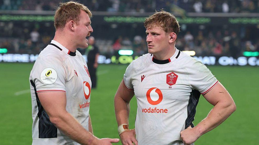 Dewi Lake (left) and Jac Morgan stand facing each other on the pitch, both are wearing white Wales kit