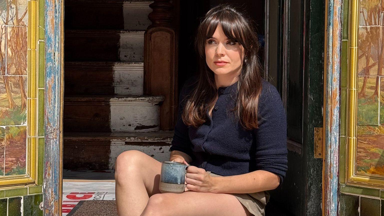 Sheri Ingram is photographed sat down in the door way of her home, which is in the process of being renovated. She is holding a blue mug which is placed on her right knee. She has long brown hair past her shoulders and is wearing a blue knitted cardigan. Steps inside her home leading upstairs can be seen behind her through the open doorway. There is Edwardian tiles on each side of the doorway, framing the photo. She is looking away from the camera and is not smiling.