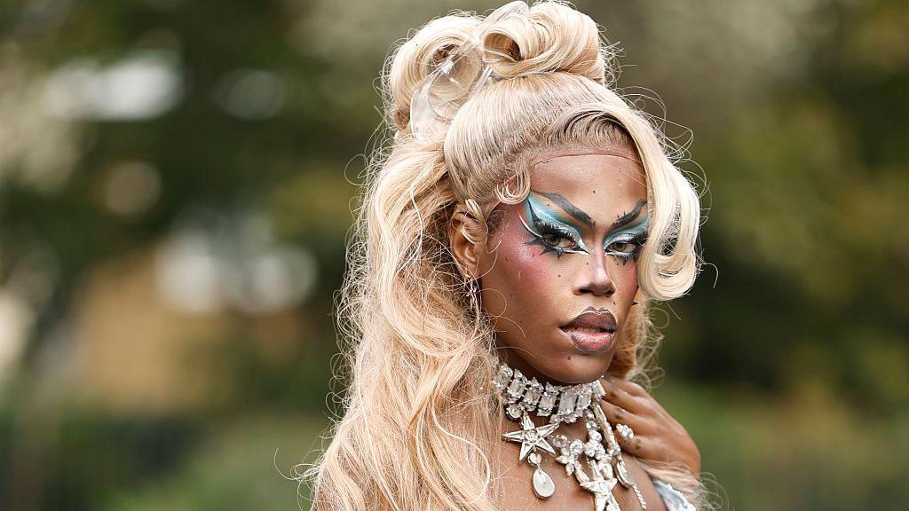 Drag queen Nyongbella, a headshot of her posing. She is wearing a blond wig in a high ponytail with a curly side fringe. She has huge blue and black eye makeup on and a chunky diamante and silver necklace.