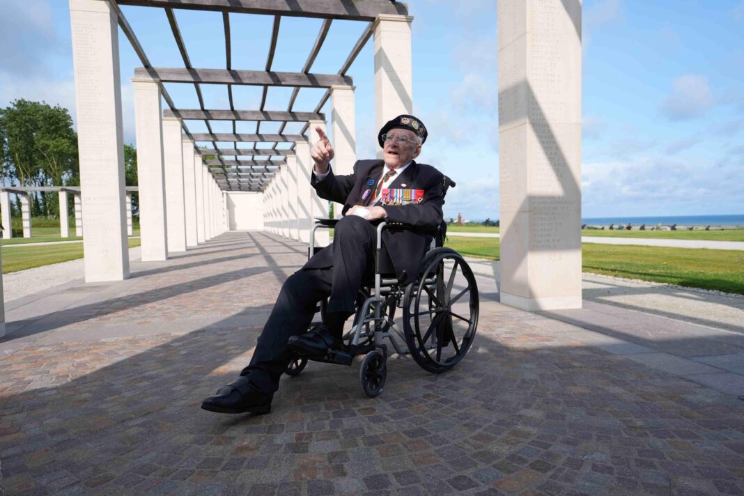 Hay in a wheelchair underneath several square arches in front of a large scale memorial pointing at something in the distance