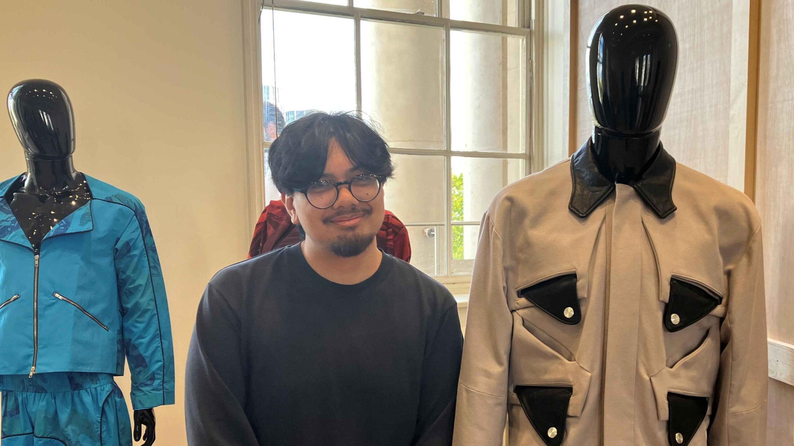 Sean stands with his fashion display behind him. He wears a blue jumper and has short black hair and dark-rimmed glasses. He stands between three mannequins, one with a beige jacket, the second with a blue sports style co-ord and part of the third can be seen, which is a red patterned jacket.