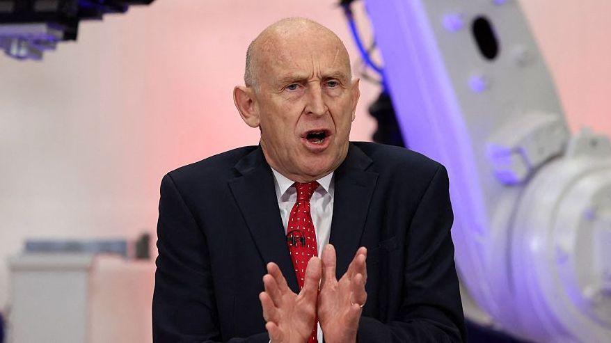 Chancellor John Healey speaking in Coventry in front of purple and grey coloured equipment. He is wearing a dark suit, white short and red tie