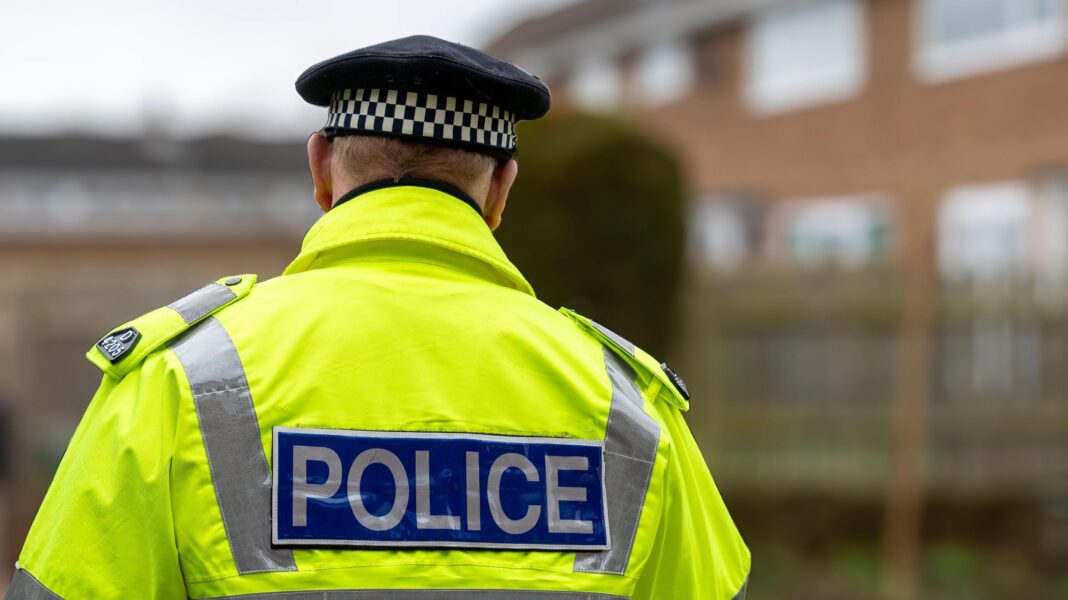 Police forces failing investigation basics, says inspectorate report A policeman in a high-vis uniform jacket with 'POLICE' written on the back is facing away from the camera. He is wearing a police hat. There are buildings blurred in the background.