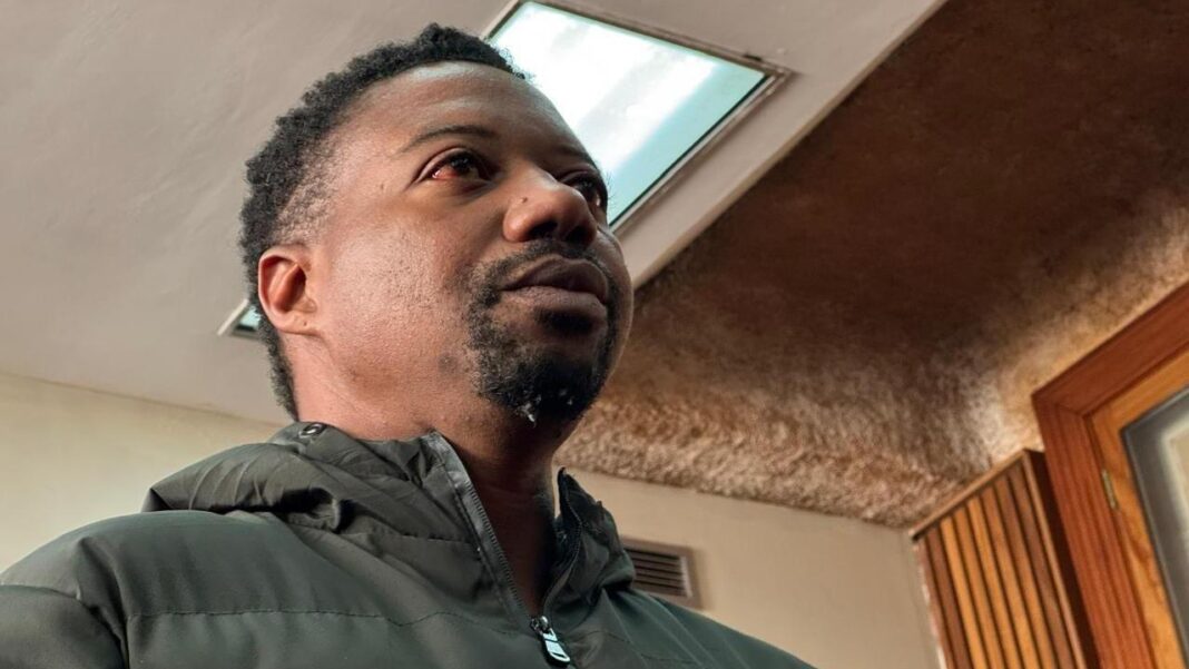 Ndodana Mkhanyisi Tshuma: The head and shoulders of a man with dark hair and a dark beard. He is wearing a dark green coat with a hood.